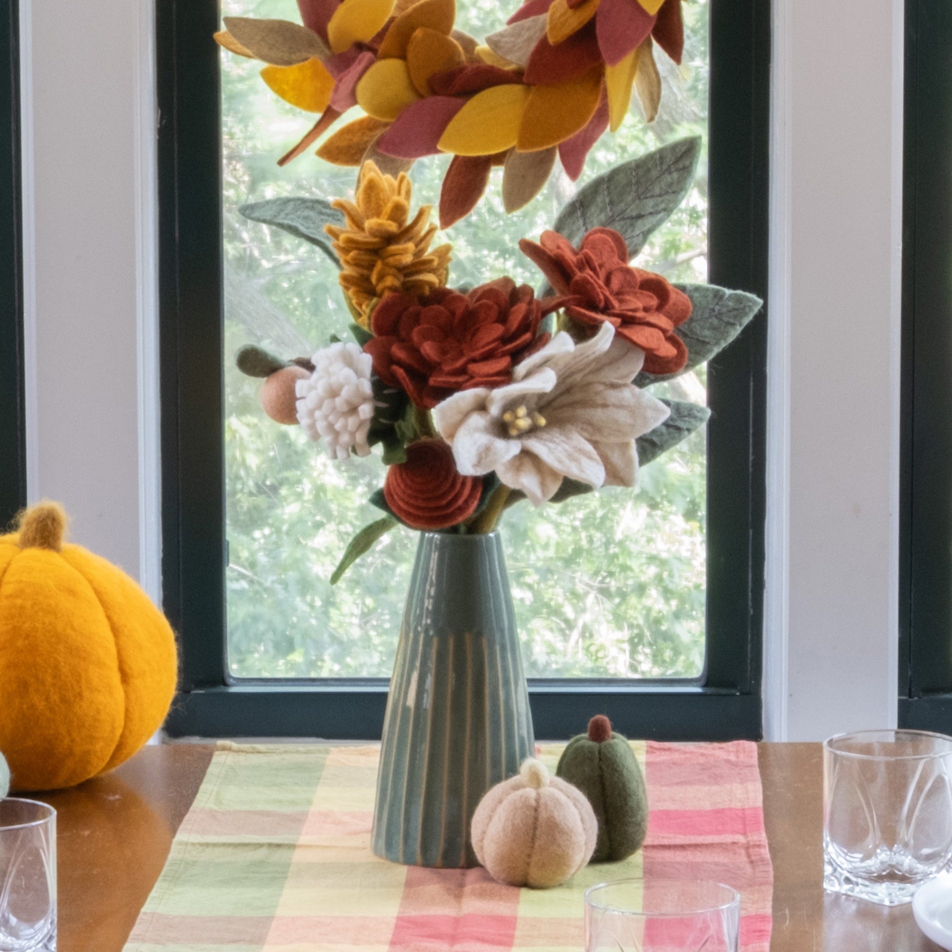 Dining room table set for a meal with autumn decorations, including pumpkins and floral arrangements, in front of a large window.